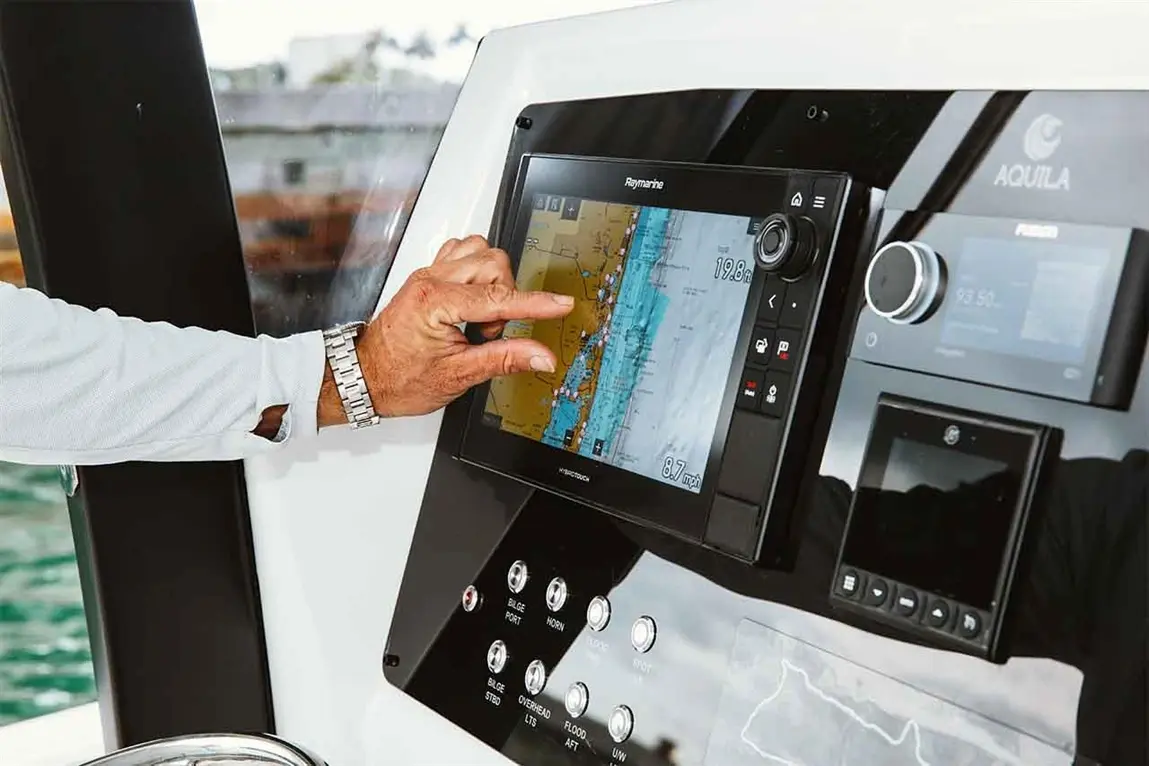 A person interacts with a Raymarine navigation display on a boat, showing maps and speed information, including 19.8 mph.